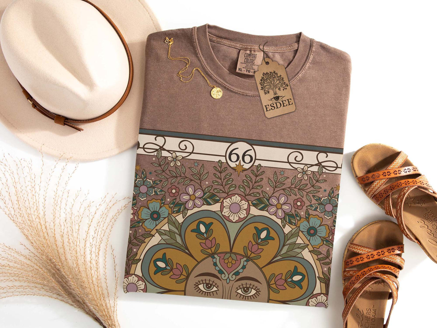 Folded brown t-shirt with decorative design and '66' on a white surface, surrounded by a hat and sandals.