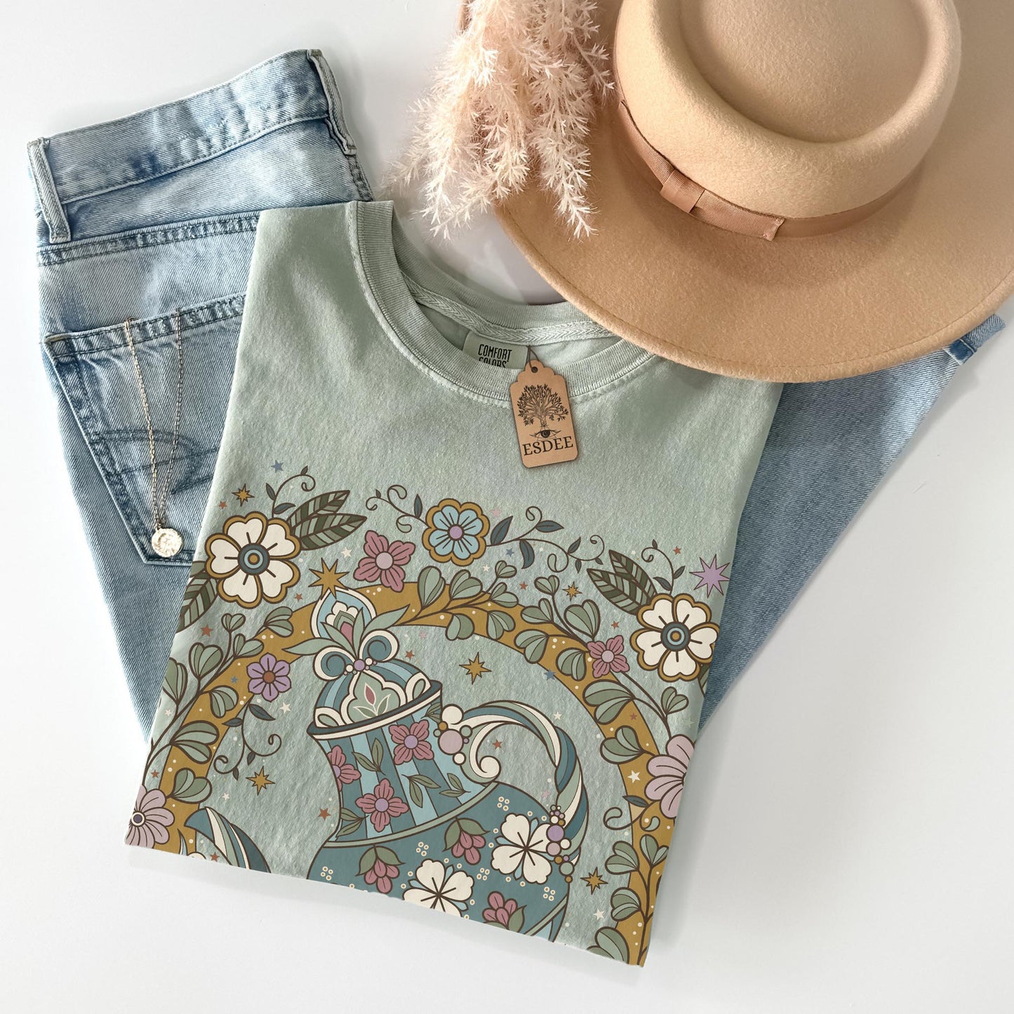 Light blue t-shirt with floral teapot design, denim jeans, and beige hat on a white background