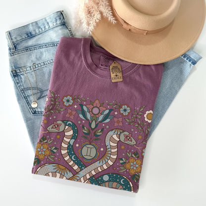 Purple t-shirt with snake and floral design on a white surface with jeans and a hat.