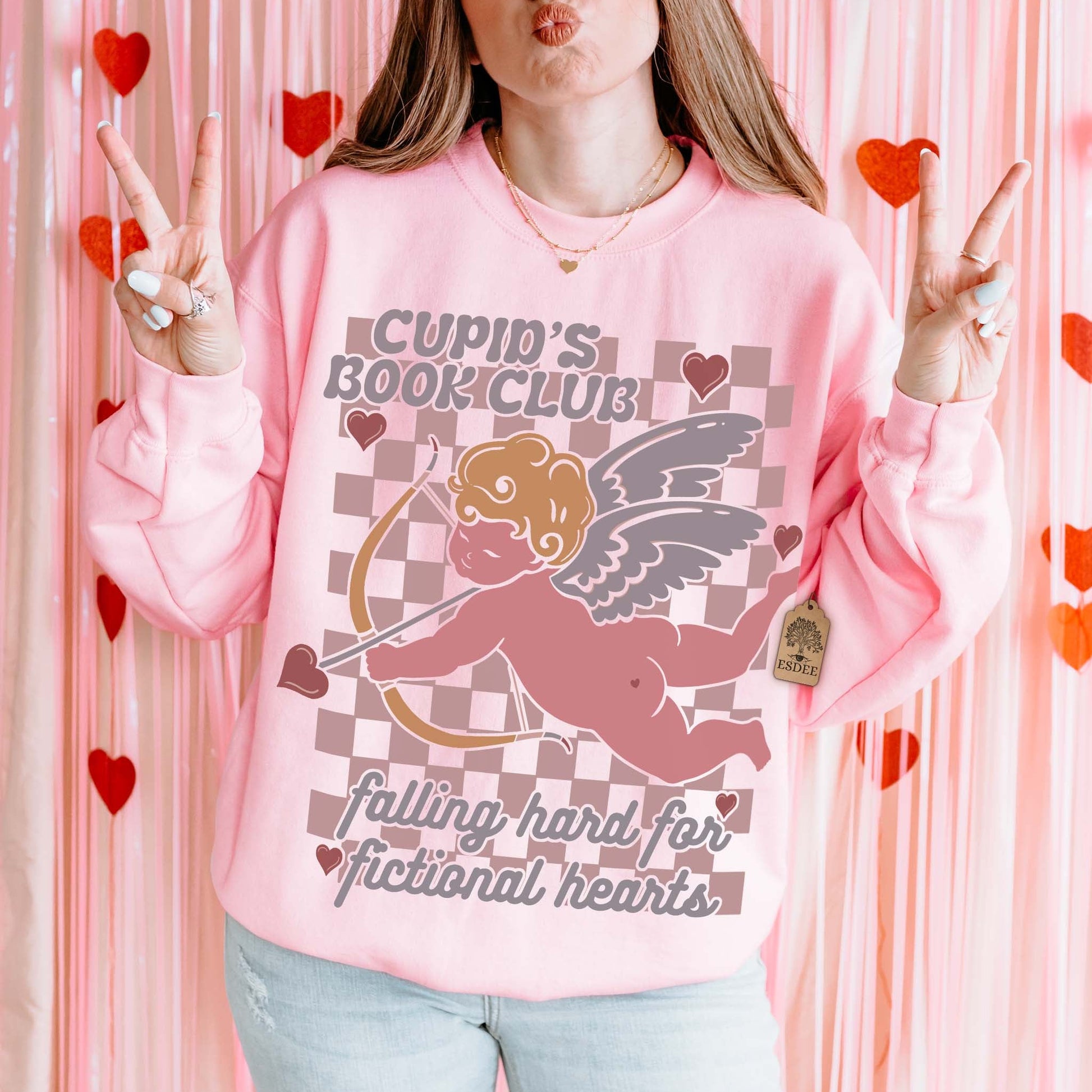 Person wearing a pink sweatshirt with 'Cupid's Book Club' design, making peace signs against a pink heart-patterned background.