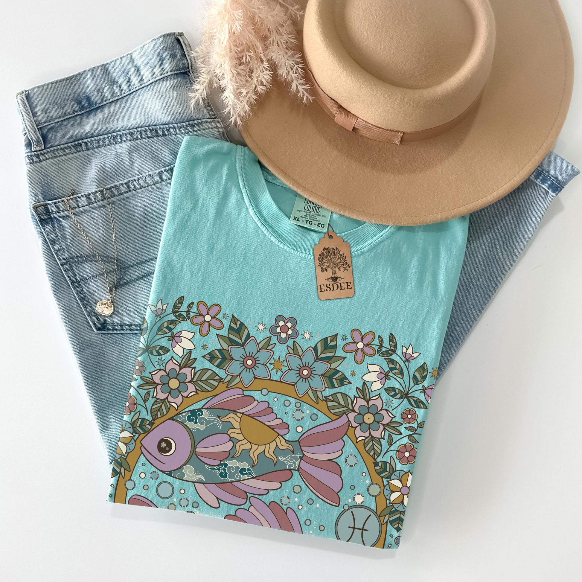Folded teal t-shirt with floral and fish design, denim shorts, and a beige hat on a white background