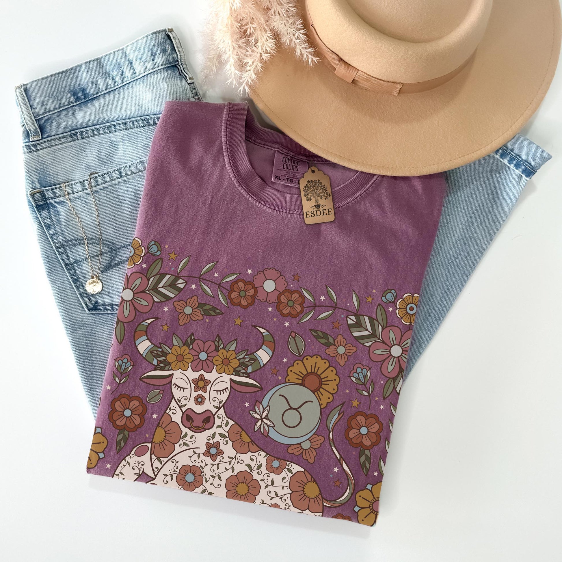 Purple t-shirt with floral and bull design on a white surface with jeans and a hat.