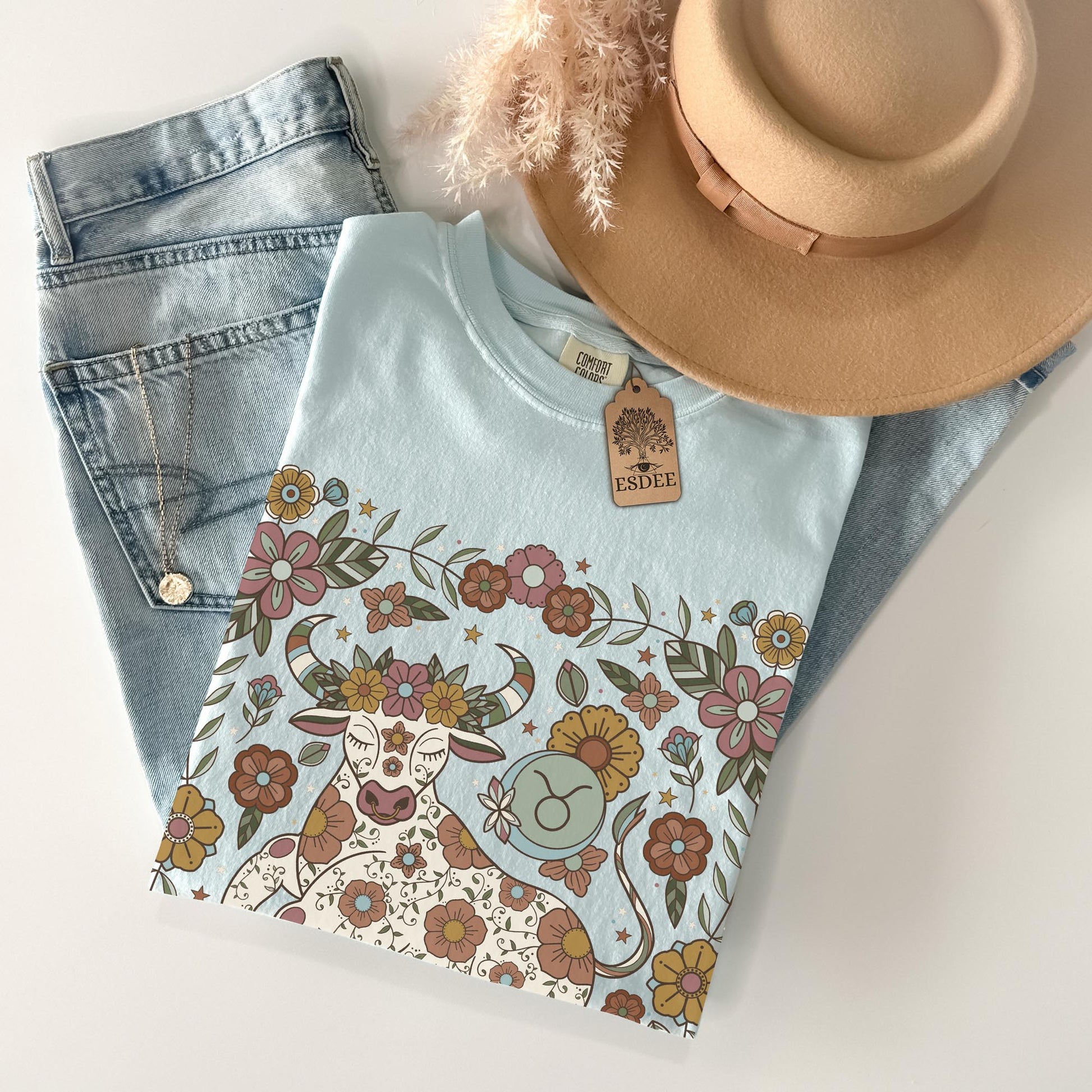 Light blue t-shirt with floral and cow design, denim jeans, and beige hat on a white surface