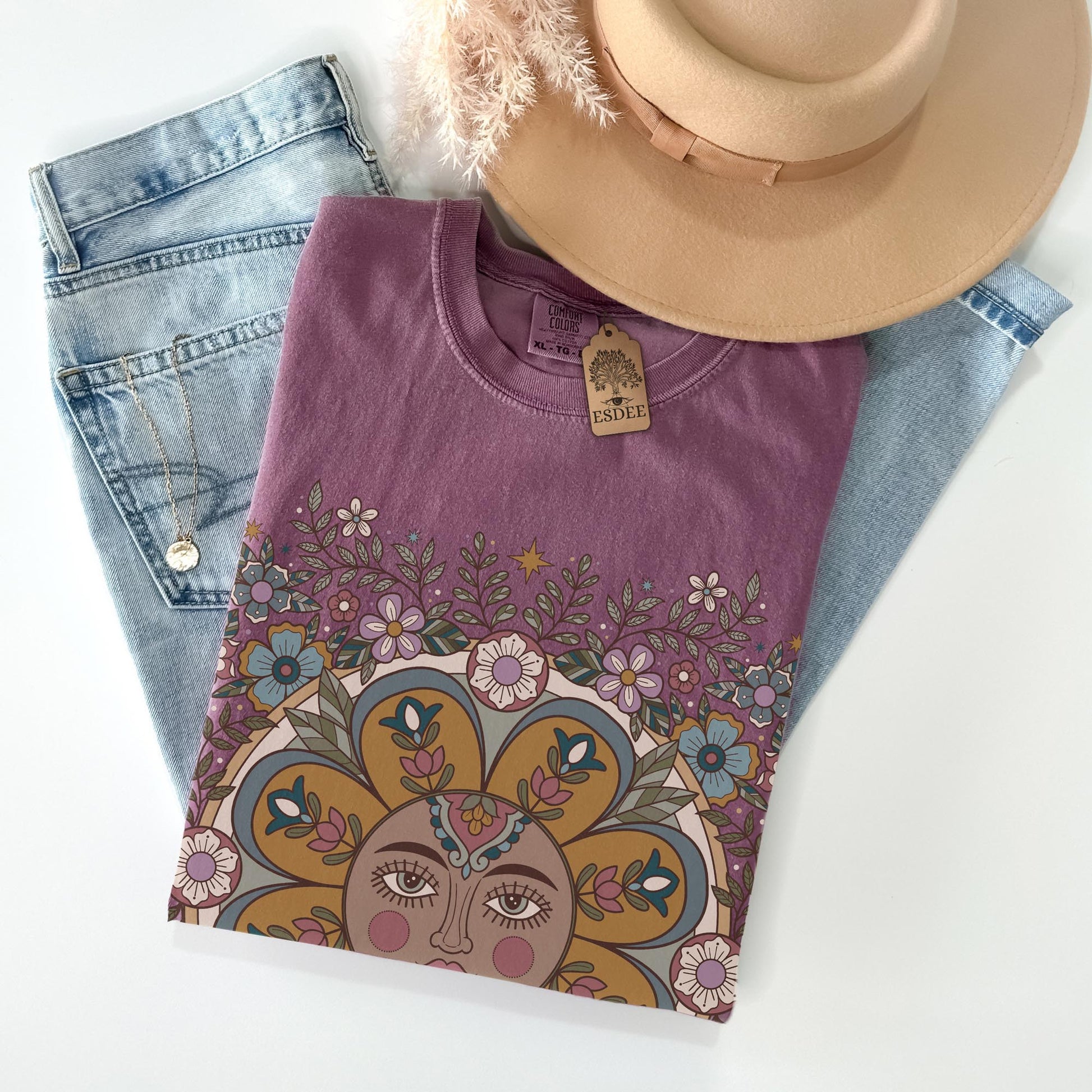 Purple t-shirt with a colorful design, placed on denim jeans and a beige hat on a white background.